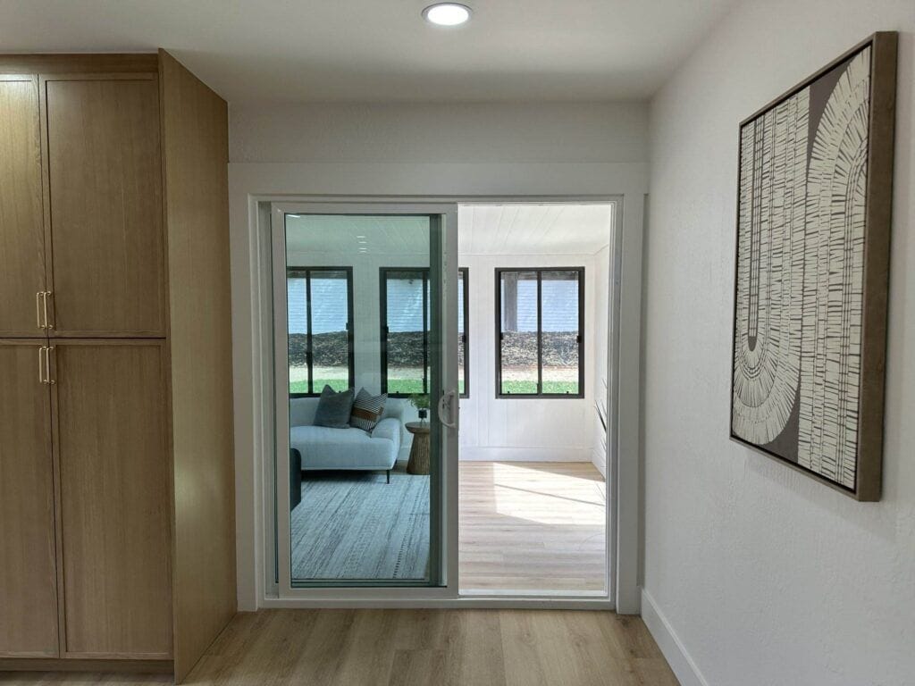 A wooden floor hallway with built-in cabinets on the left, a large abstract painting on the right, and a sliding glass door leading to a sunroom with a sofa and multiple windows.