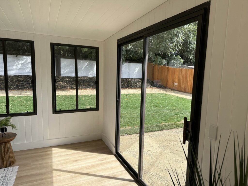 A sunlit enclosed patio with white walls, large black-framed windows, and a sliding glass door opening to a grassy backyard with wooden and stone fencing.