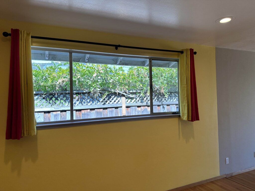 A large window with curtains partially drawn in a room with yellow walls. Outside, there is a wooden fence and green plants visible.