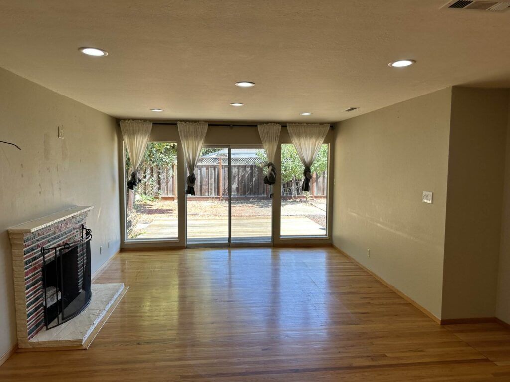 A vacant living room with hardwood floors, a fireplace on the left, and sliding glass doors with curtains leading to a backyard. Recessed ceiling lights are turned on.