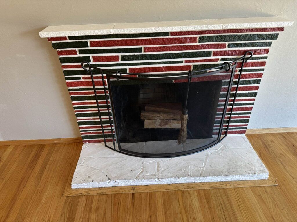 Brick fireplace with a black metal screen, containing stacked firewood inside. The bricks are horizontally layered in red, white, and black colors. The hearth has a white stone surface.