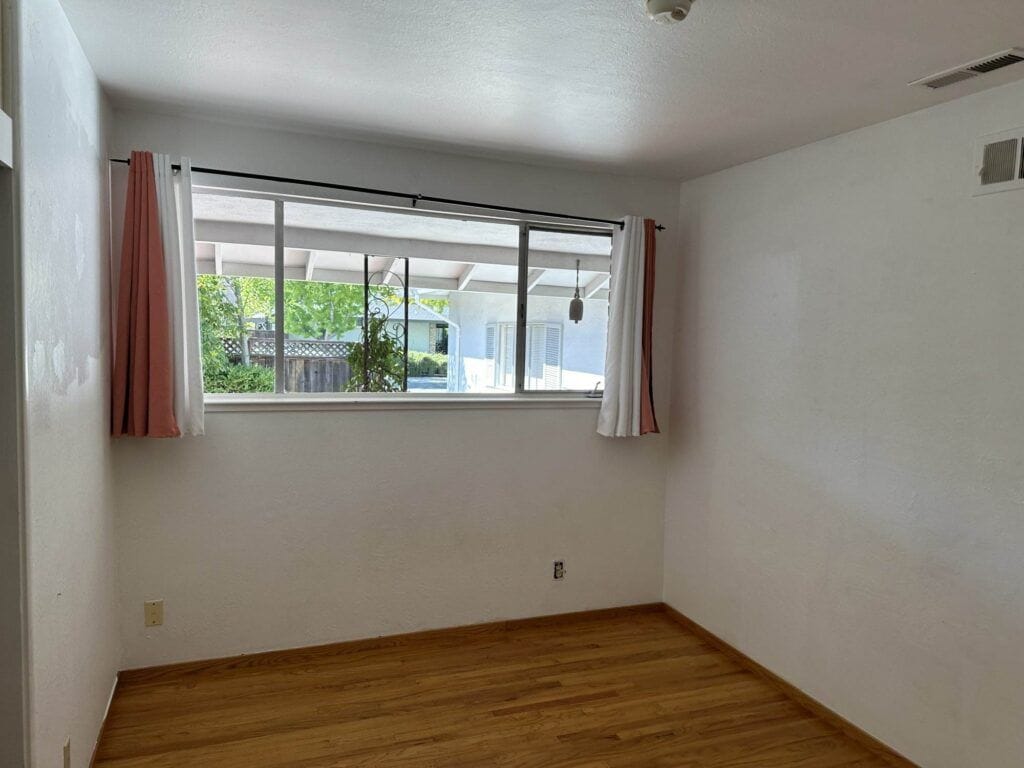 A small empty room with light-colored hardwood floors, white walls, and a large window with white and pink curtains, looking out to a covered patio and greenery.