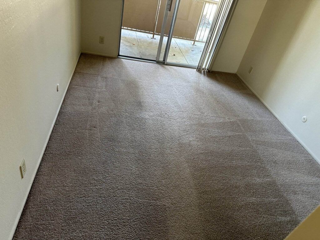 Empty room with beige carpet, beige walls, and a sliding glass door leading to a small balcony. Vertical blinds are partially open, allowing sunlight to enter. No furniture is present.