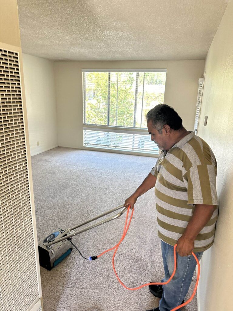 A man in a striped shirt operates a carpet cleaning machine in a light-filled, empty room with a large window.