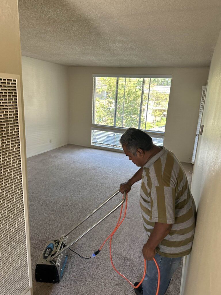 A man operates a carpet cleaning machine in an empty room with beige walls and large windows.