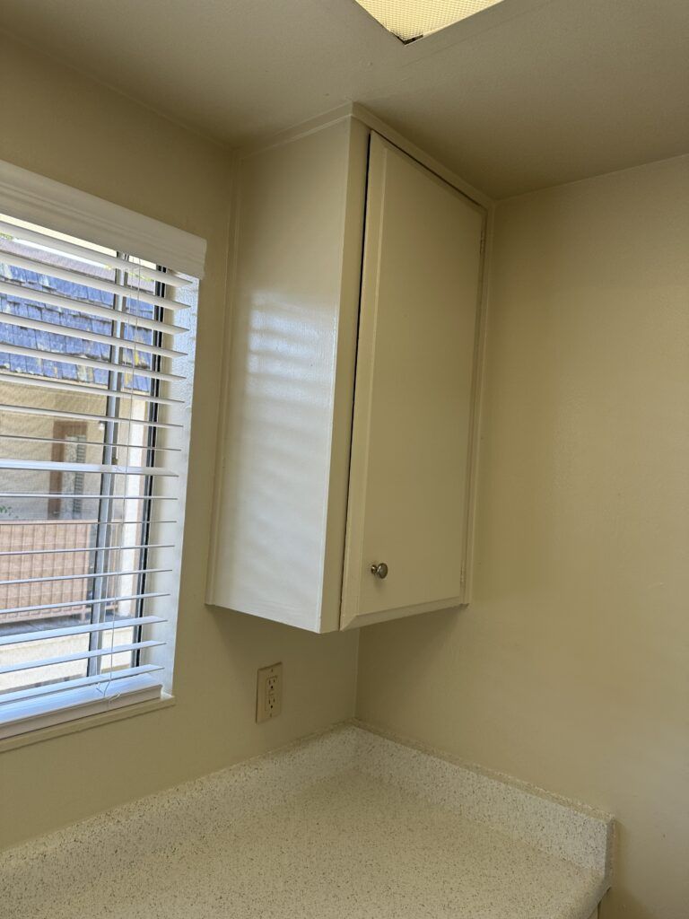 A kitchen corner with a small upper cabinet, countertop, and window with blinds. The wall and cabinet are painted light yellow. An electrical outlet is visible below the cabinet.