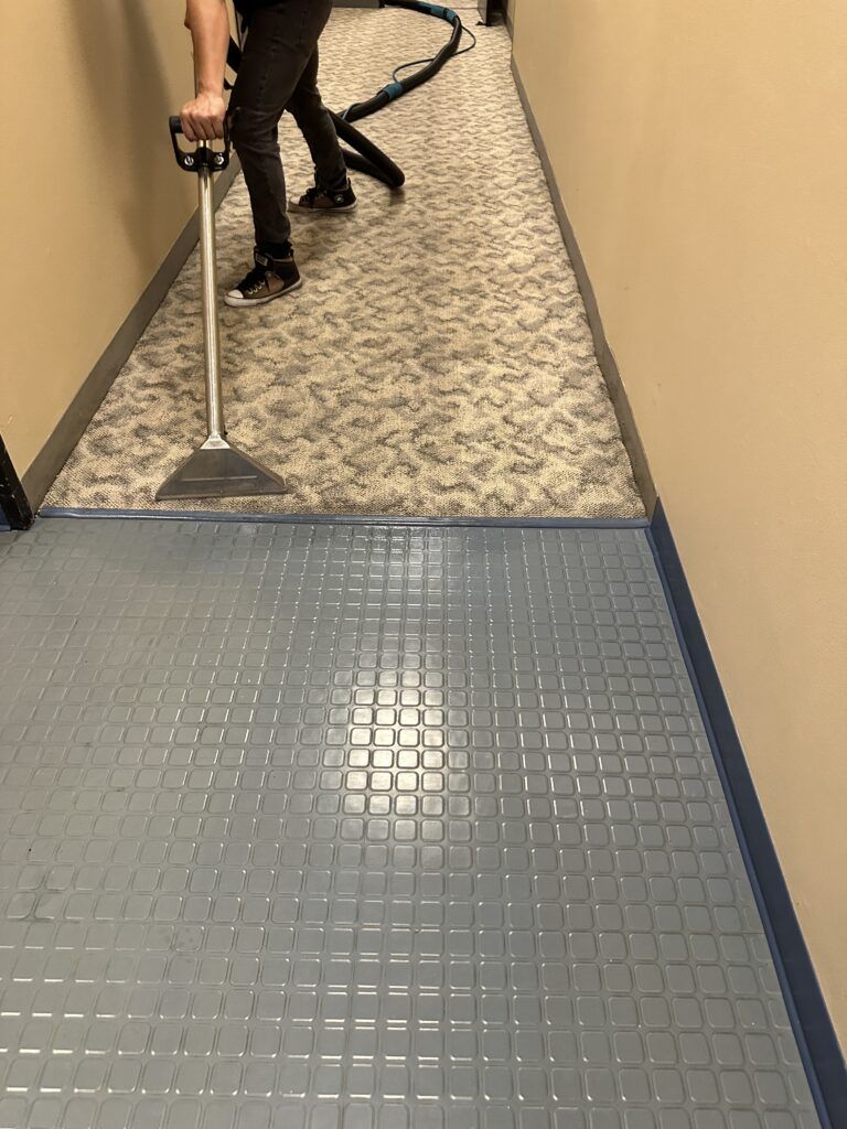 Person using a carpet cleaning machine on a patterned carpet in a hallway. Blue-tiled floor area in the foreground.