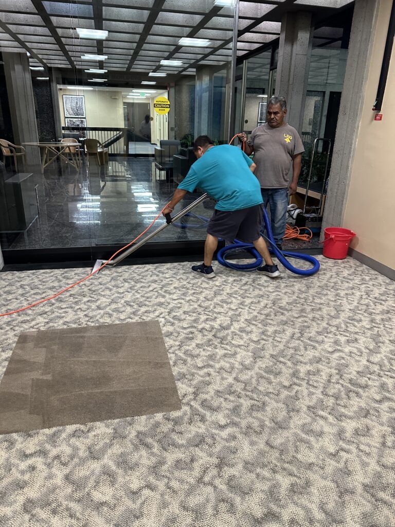 Two men are working indoors; one is holding a blue hose and the other is managing cables. The area features glass walls, marble flooring in the background, and carpeted flooring in the foreground.