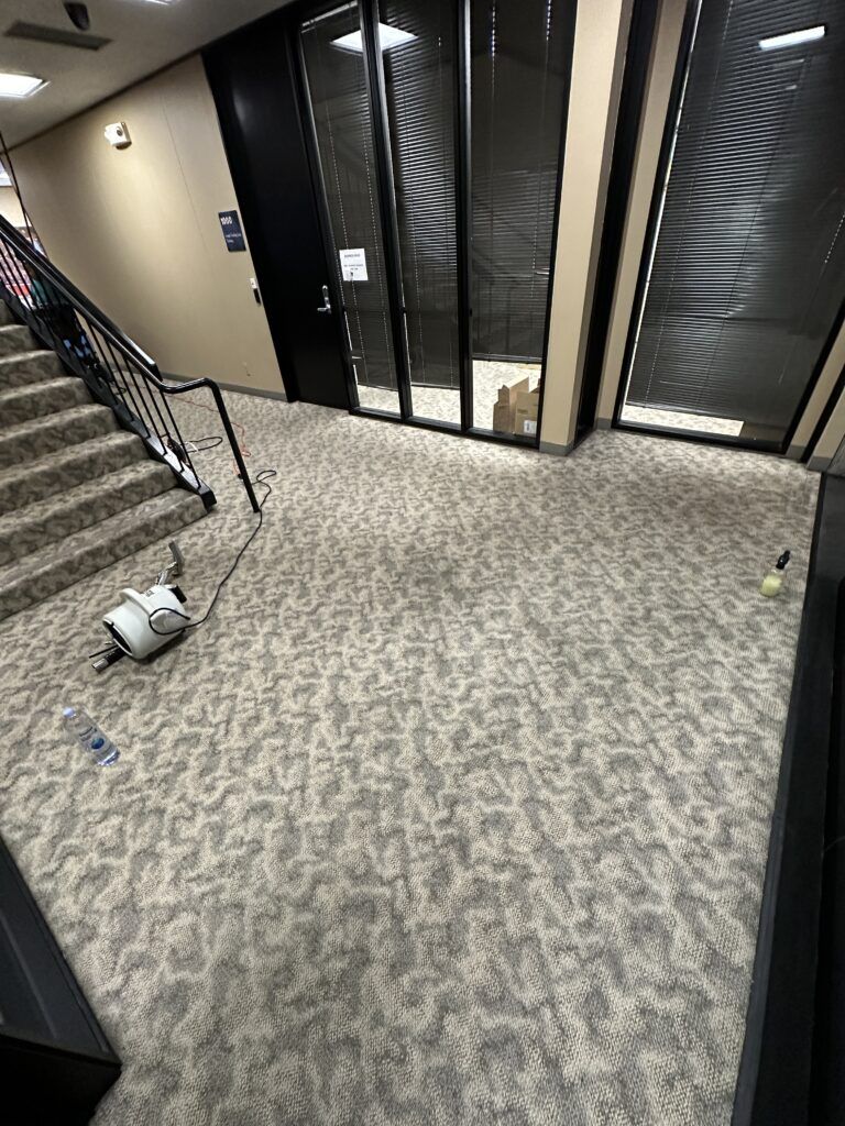 A carpeted hallway with a cleaning vacuum, water bottle, and cleaning utensil on the floor next to a staircase. Glass doors with blinds are visible in the background.