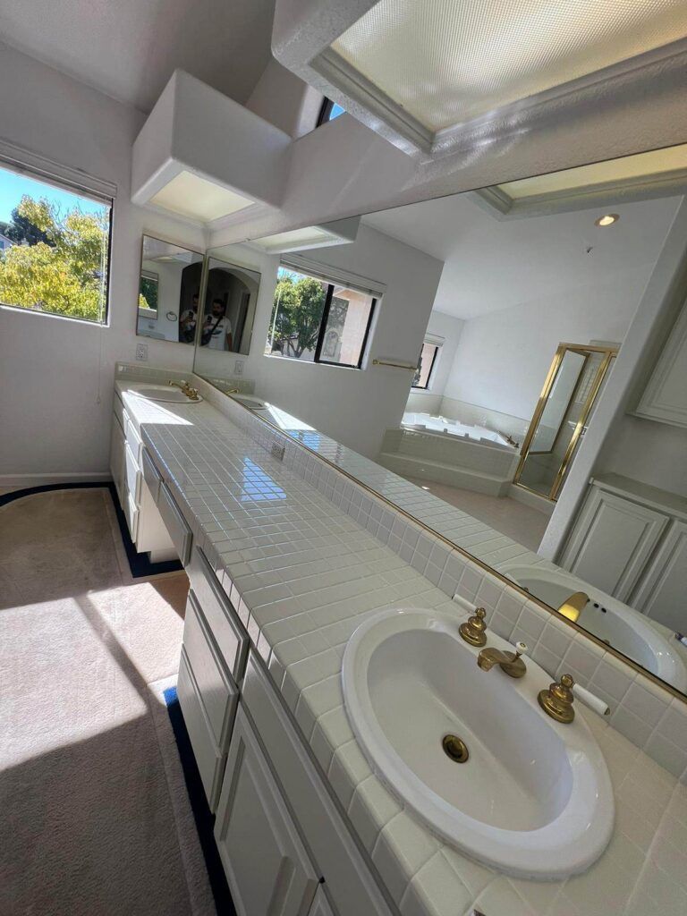 A bathroom featuring a long countertop with dual sinks, brass fixtures, large mirrors, a window, a shower with glass doors, and a soaking tub. Sunlight streams in through a window.