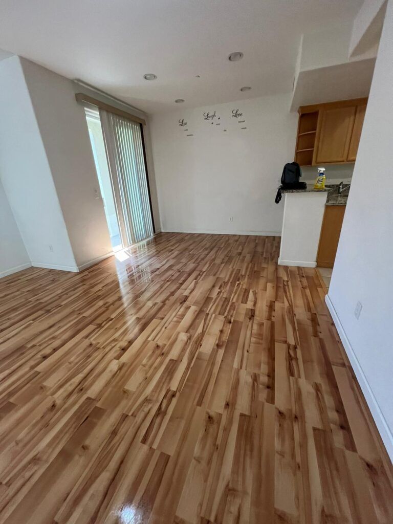 A vacant room with polished wooden floors, a sliding glass door leading outside, light-colored walls, and a partially visible kitchen area with wooden cabinetry and a backpack on the counter.