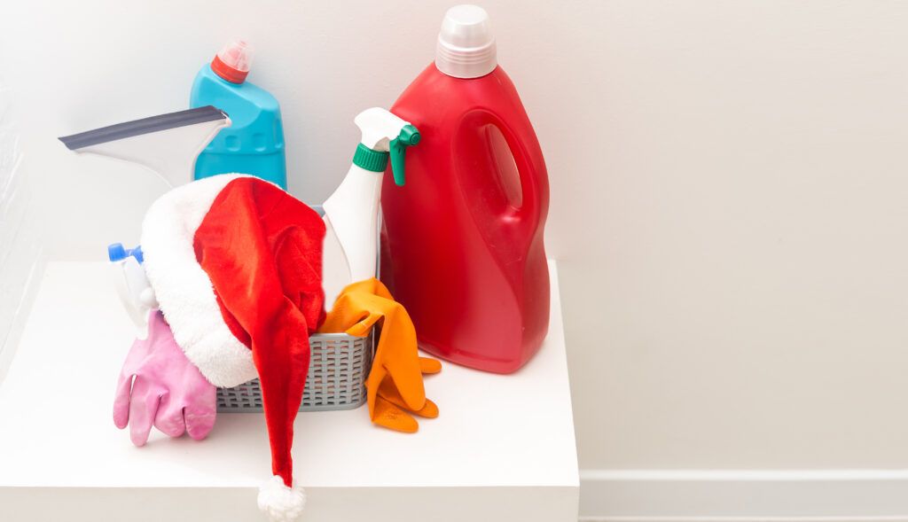 A basket holds cleaning supplies, including a red detergent bottle, spray cleaner, gloves, squeegee, and a Santa hat on a white surface against a plain wall—perfect for anyone in need of Tailored Cleaning Services in Santa Clara Home.