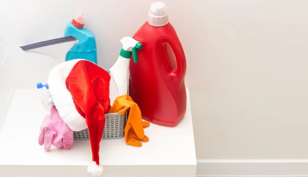 A basket holds cleaning supplies, including a red detergent bottle, spray cleaner, gloves, squeegee, and a Santa hat on a white surface against a plain wall—perfect for anyone in need of Tailored Cleaning Services in Santa Clara Home.