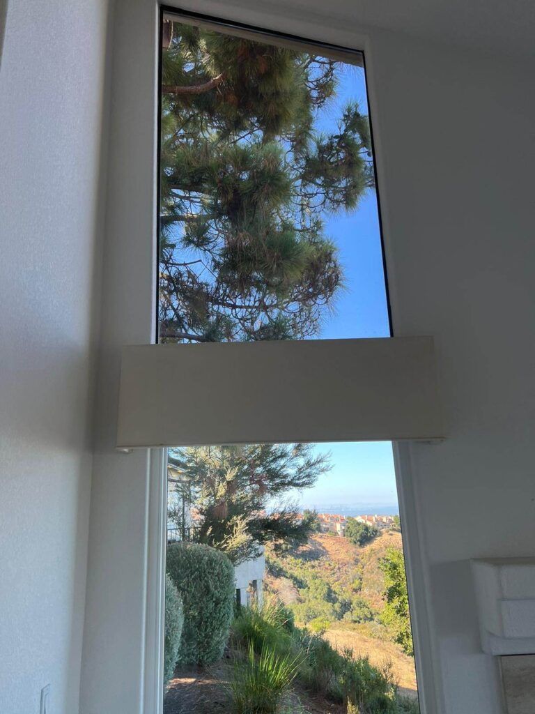 A large window with a wide wooden frame provides a view of a pine tree, bushes, and distant hills under a clear blue sky.