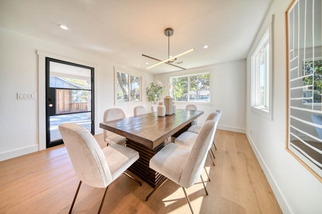 A modern dining room with a wooden table, eight upholstered chairs, a potted plant, and a large window allowing natural light. There is a door leading outside in the background.
