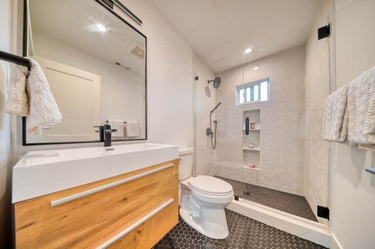 Modern bathroom with a wooden vanity and white sink, wall-mounted mirror, towel rack, toilet, and walk-in shower with glass door and hexagonal floor tiles.