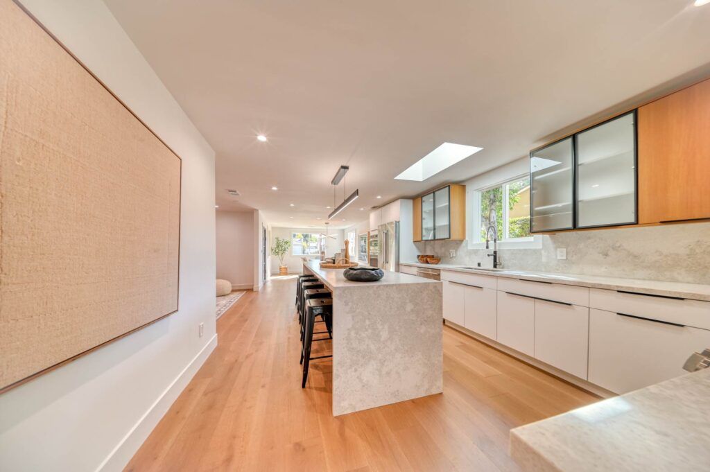 A modern kitchen with light wood flooring, white cabinets, a long island with bar stools, and minimalist decor. The room features recessed lighting and a large window for natural light.