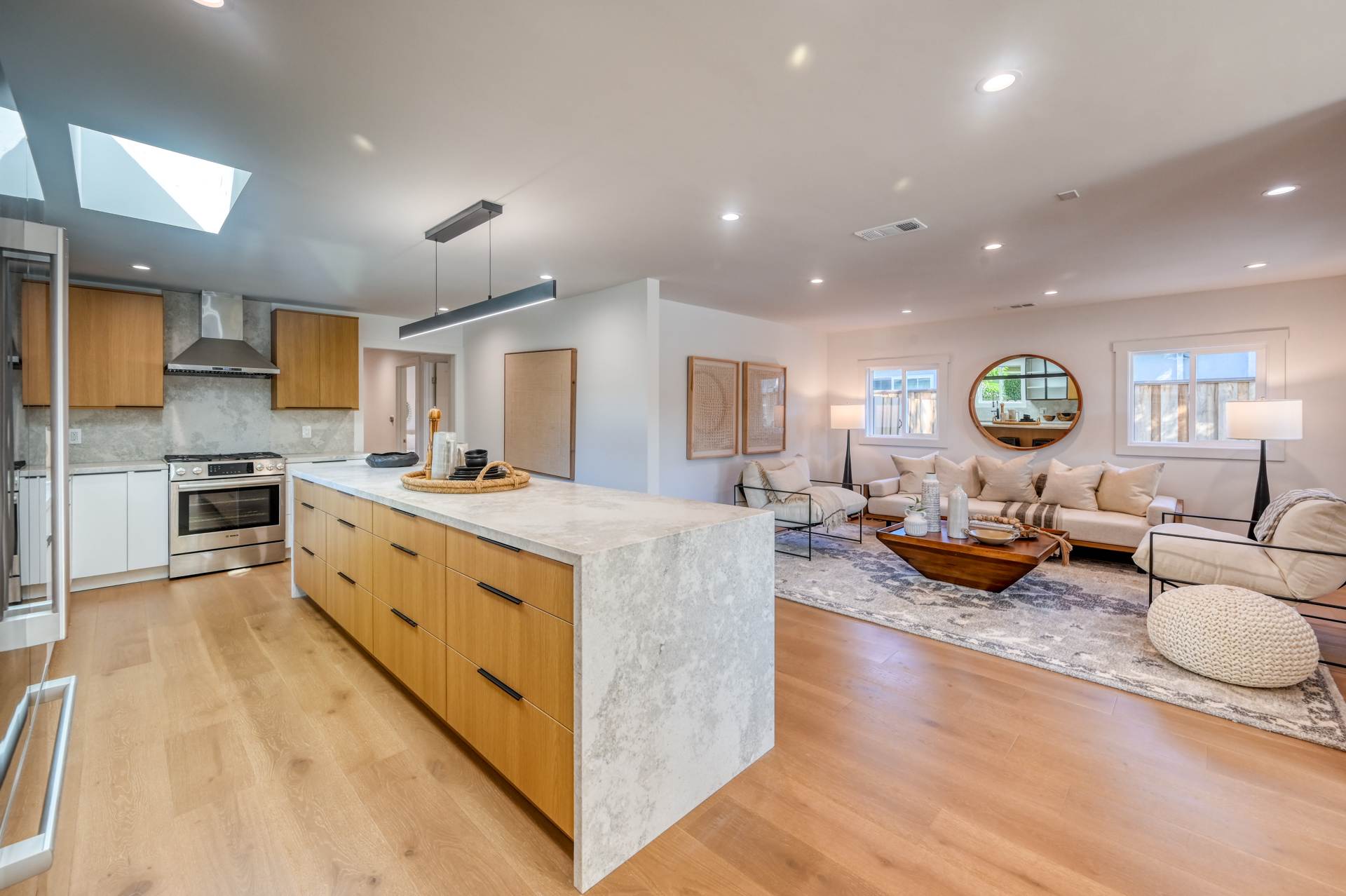 Modern open-concept living space with a kitchen island, light wooden flooring, minimalist furnishings, and recessed lighting. Kitchen has stainless steel appliances, and the living area features a large sofa.