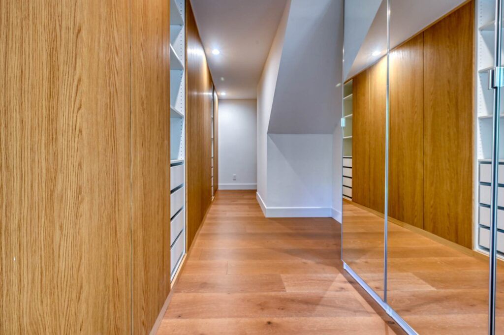 A hallway with wooden flooring and walls lined with built-in shelves. One side features a large mirrored door. The space is well-lit.