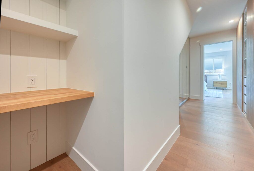 A hallway with light wood flooring, white walls, and recessed lighting leads to a room with a bed. A wooden ledge and electrical outlets are visible on the left.