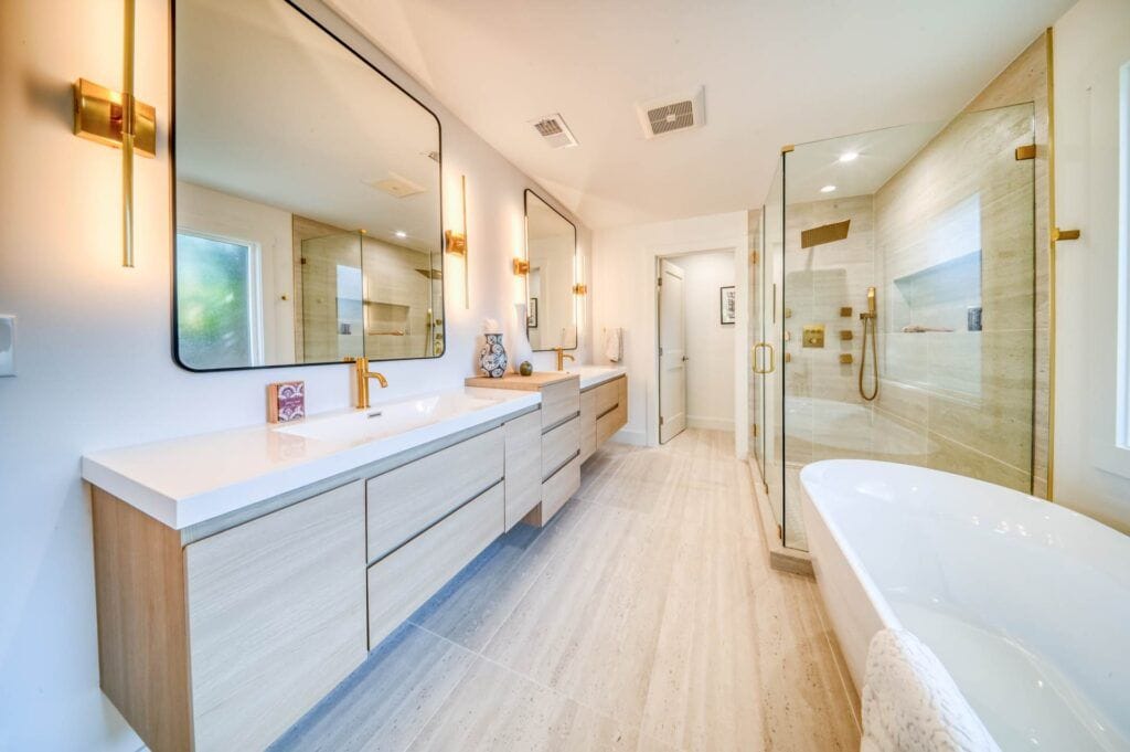 Modern bathroom with double sinks, large mirrors, wooden cabinets, and a glass-enclosed shower. The room is well-lit, featuring a freestanding bathtub and minimalist design elements.