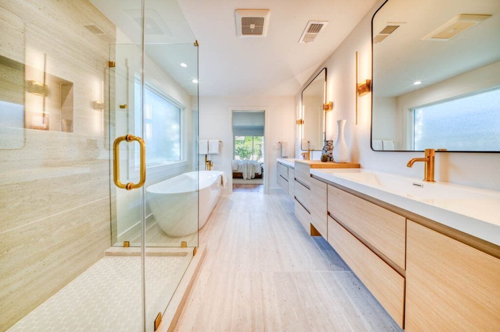 Modern bathroom with a glass-enclosed shower, freestanding tub, double vanity, and large windows, featuring light wood cabinetry and gold hardware accents.