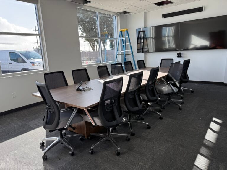 A modern conference room with a long table, ten office chairs, a large wall-mounted screen, a ladder, and sunlight streaming in—kept spotless by top-notch janitorial cleaning services.