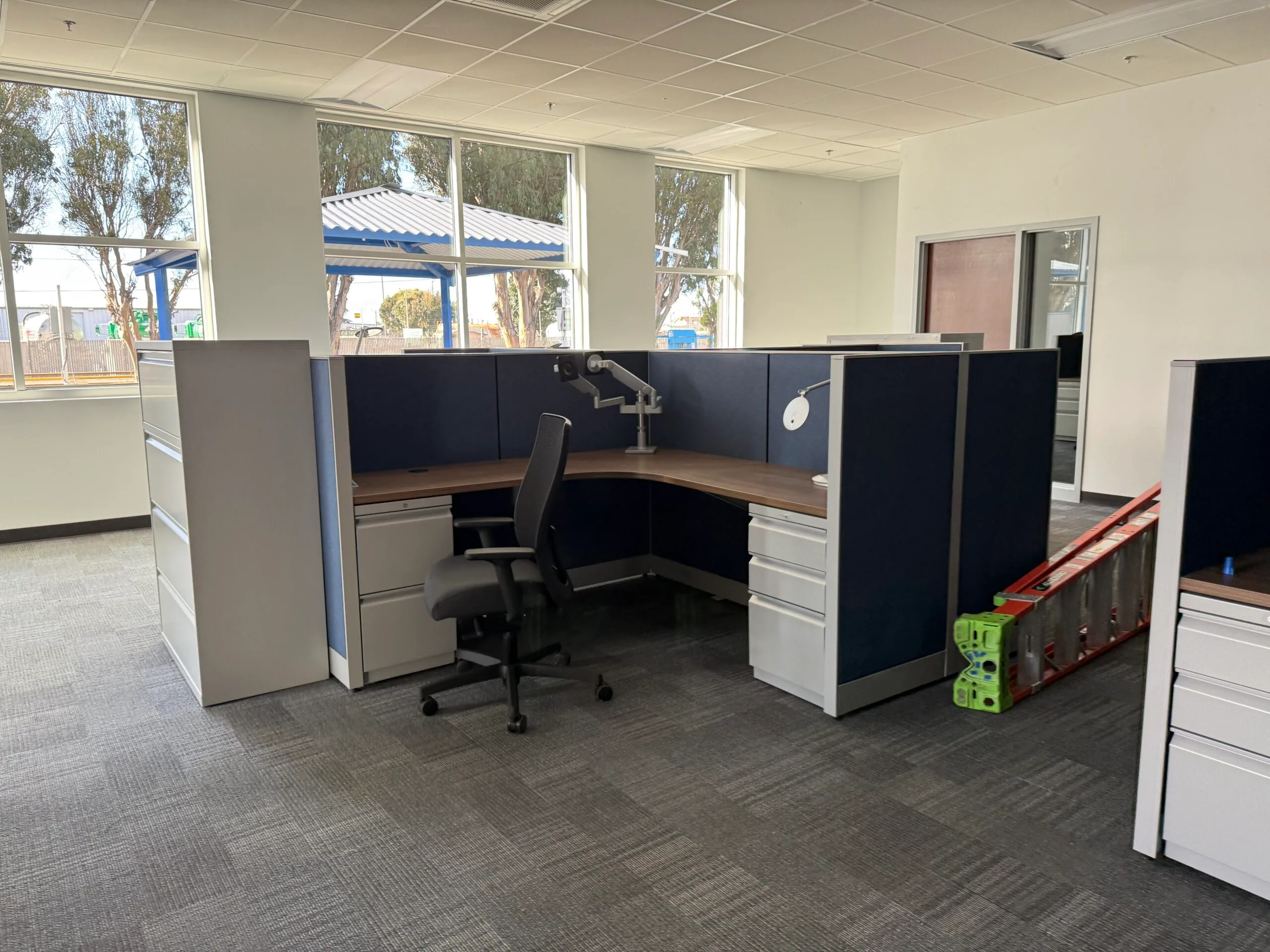 An empty office cubicle with an office chair, desks, file cabinets, and a monitor arm—ready for janitorial cleaning services. A ladder and step stool are visible in the background. Large windows let in natural light.
