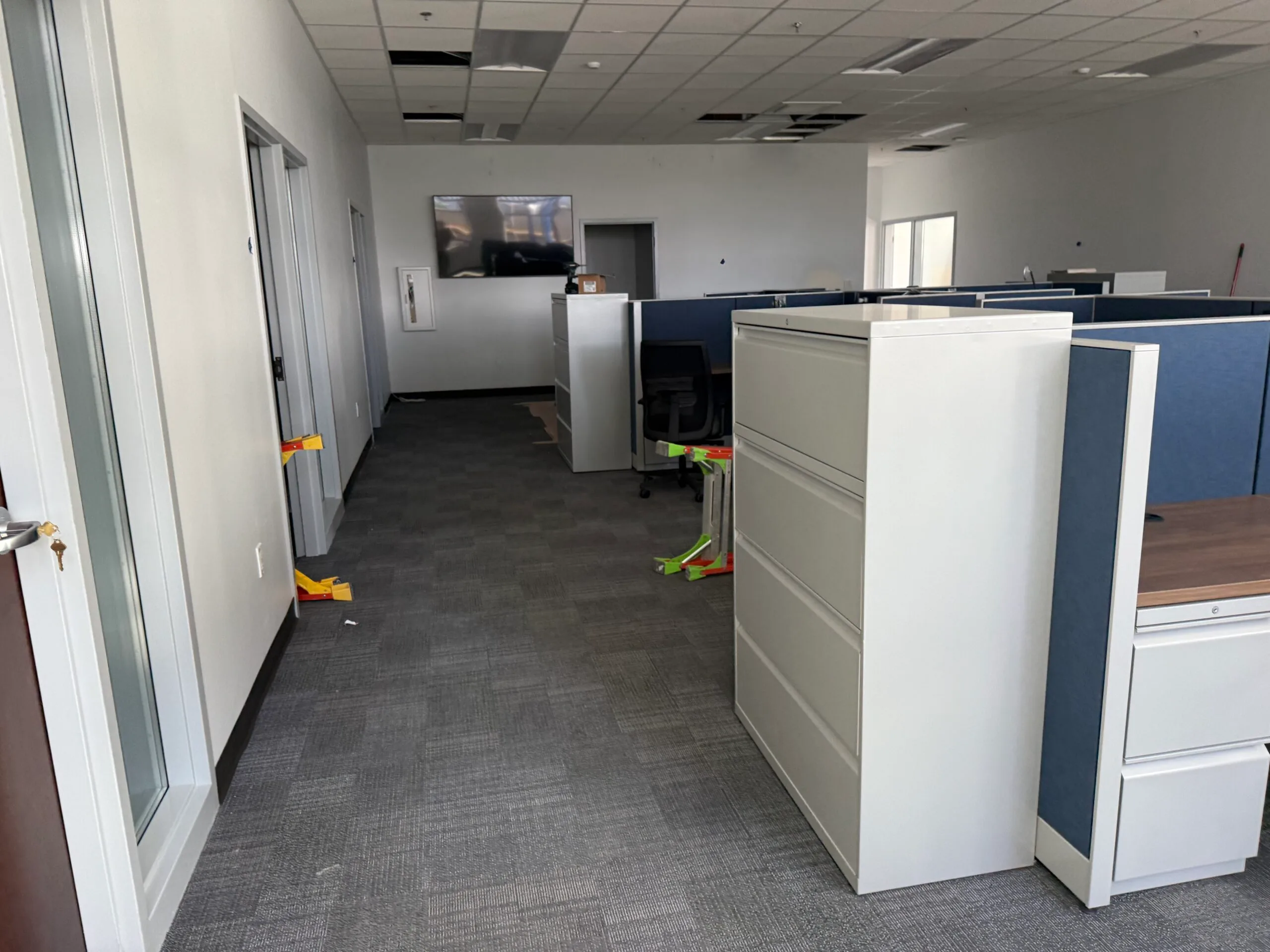 A modern office space with cubicles, filing cabinets, and office chairs; ceiling tiles are missing and straps are attached to door handles, indicating recent Janitorial Cleaning Services.