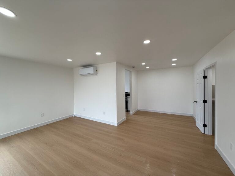 A spacious, empty room in San Jose with light wood flooring, white walls, recessed ceiling lights, a wall-mounted air conditioner, and a white door—spotless after post-construction cleaning.