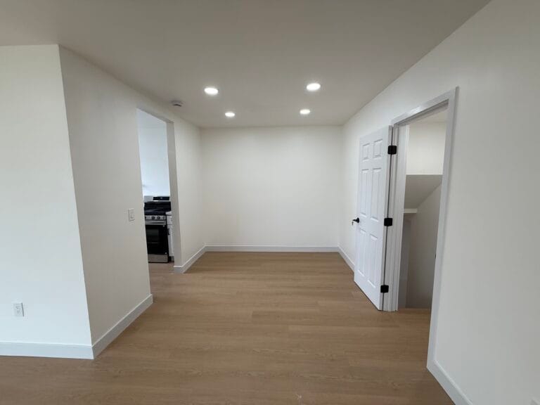 A small, empty room with wood floors, white walls, and recessed ceiling lights—perfectly refreshed after-remodel cleaning—features doors leading to a kitchen area and another room in San Jose.