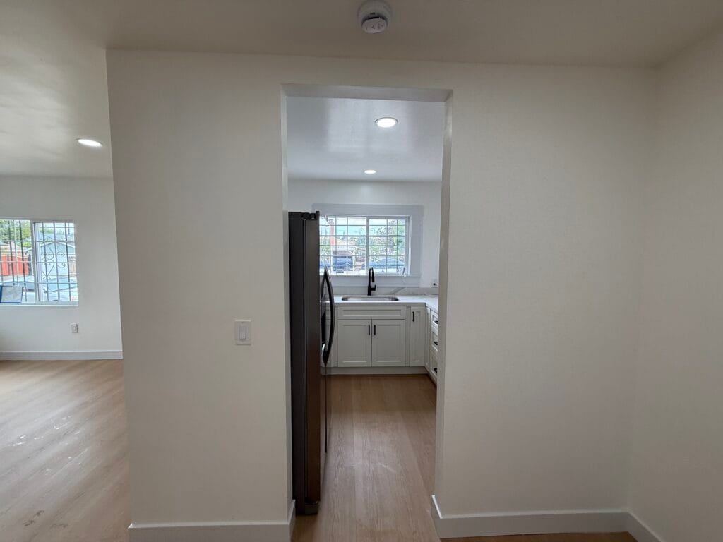View of a small kitchen in San Jose with white cabinets, stainless steel refrigerator, sink under double windows, and wood flooring—sparkling after post-construction cleaning—seen from an adjacent room.