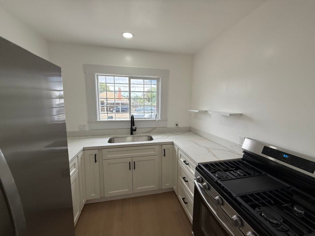 Modern kitchen in San Jose with white cabinets, stainless steel appliances, a single window above the sink, and open shelves on the right wall—spotless after detailed post-construction cleaning.