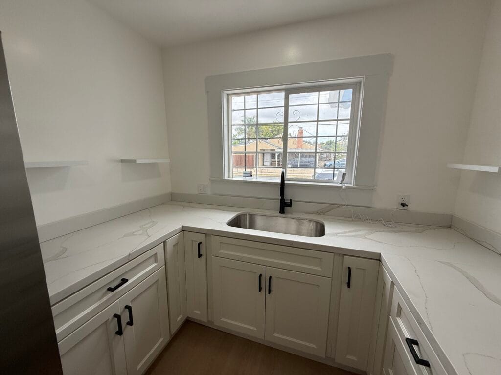 A modern San Jose kitchen with white cabinets, marble-patterned countertops, a stainless steel sink under a window, and two small white shelves—fresh and spotless after-remodel cleaning.