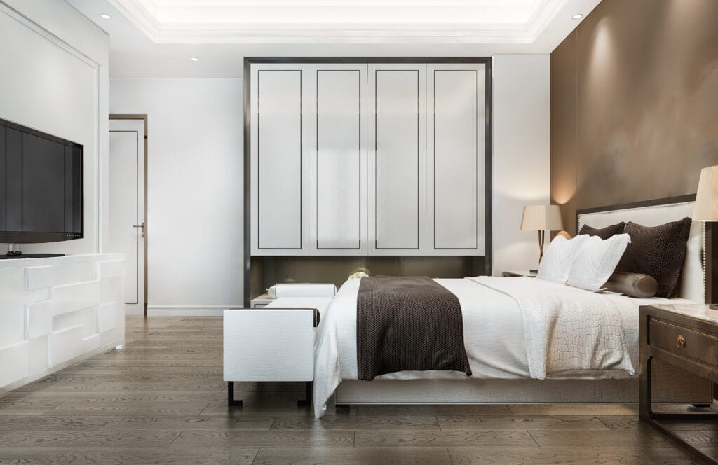 Modern bedroom with a large bed, white bedding, brown accent wall, built-in wardrobe, TV on a white stand, and wood flooring—ideal for Saratoga homeowners seeking a fresh start after post-construction cleaning. Lit by ceiling lights and a bedside lamp.