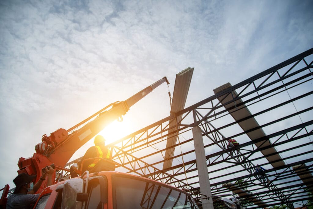 Construction workers use a crane to lift a steel beam onto a building framework under a partly cloudy Saratoga sky at sunset, preparing the site for post-construction cleaning.