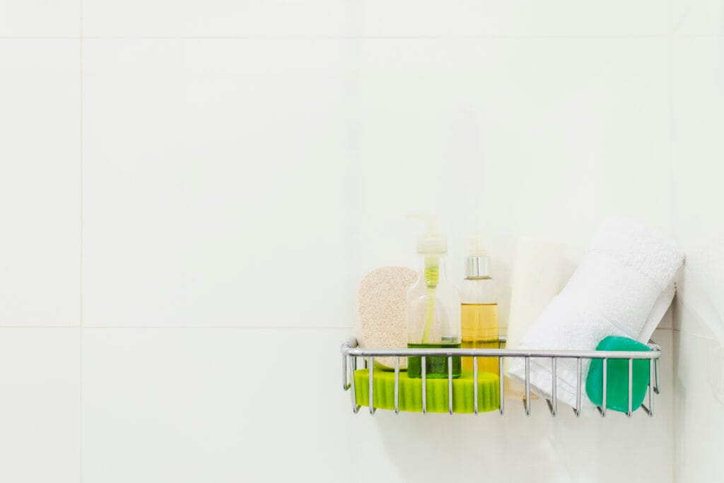 A metal shower caddy holds bottles of soap, a sponge, a loofah, a folded white towel, a rolled-up towel, and a green soap bar—perfect for homeowners focusing on bathroom deep cleaning against crisp white tiled walls.