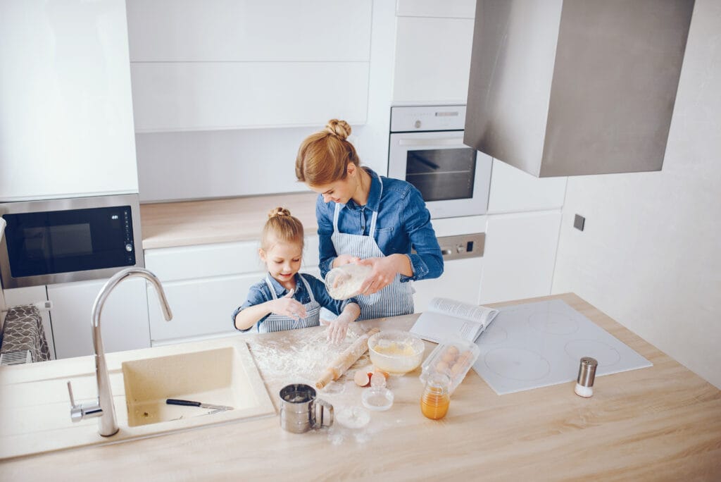 An adult and a child, both wearing aprons, bake together in a modern kitchen as they measure flour over a counter—just as meticulous as customized bathroom cleaning in Saratoga.