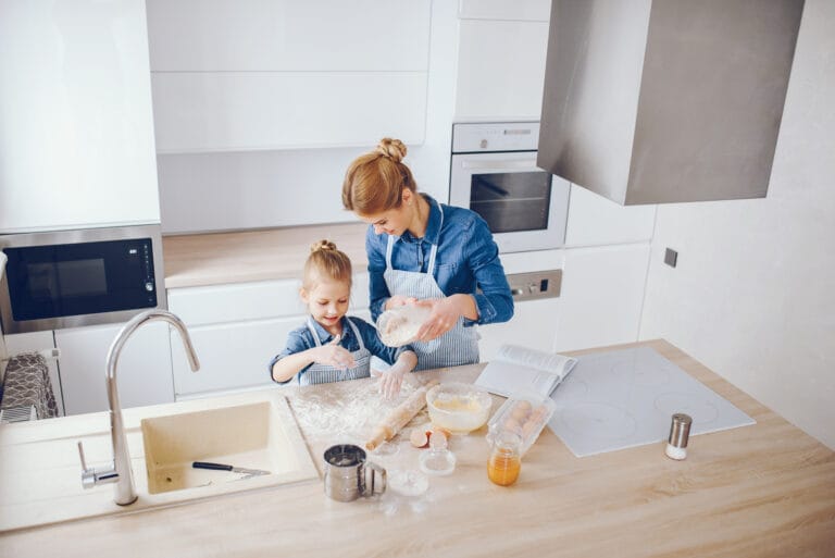 An adult and a child, both wearing aprons, bake together in a modern kitchen as they measure flour over a counter—just as meticulous as customized bathroom cleaning in Saratoga.