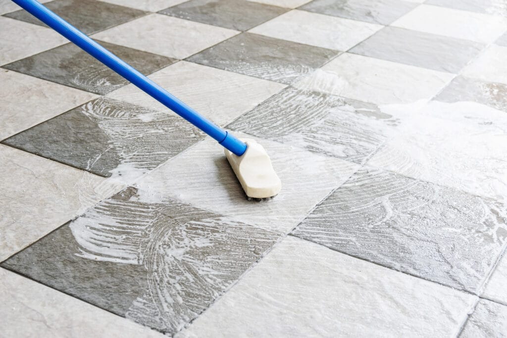 A brush with a blue handle is scrubbing soapy water on a black and white tiled floor, perfect for Property Managers seeking thorough Move-Out Cleaning Los Gatos solutions.