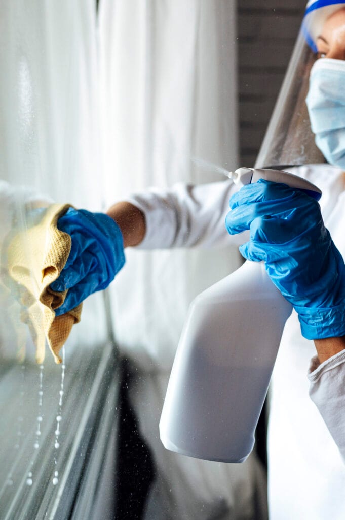 A person wearing gloves, mask, and face shield sprays cleaning solution and wipes a glass window with a yellow cloth, demonstrating the thorough care provided by Los Gatos deep cleaning services for homeowners.