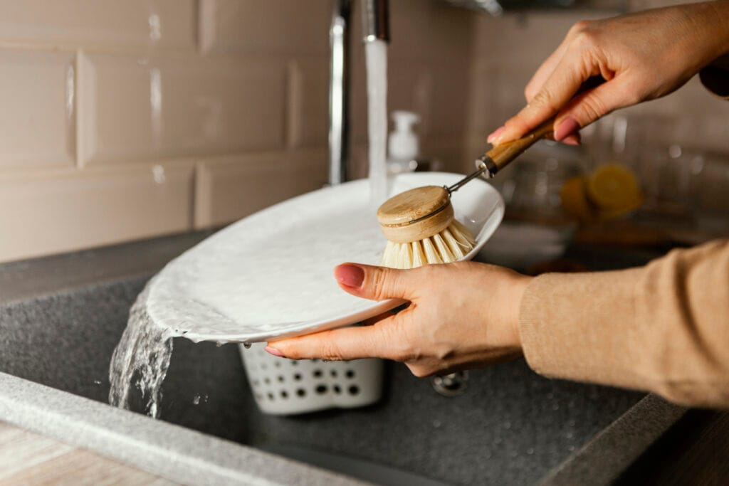 A homeowner practices Kitchen-Only Cleaning as they scrub a white plate with a brush under running water in their kitchen sink, following Campbell’s trusted cleaning tips.