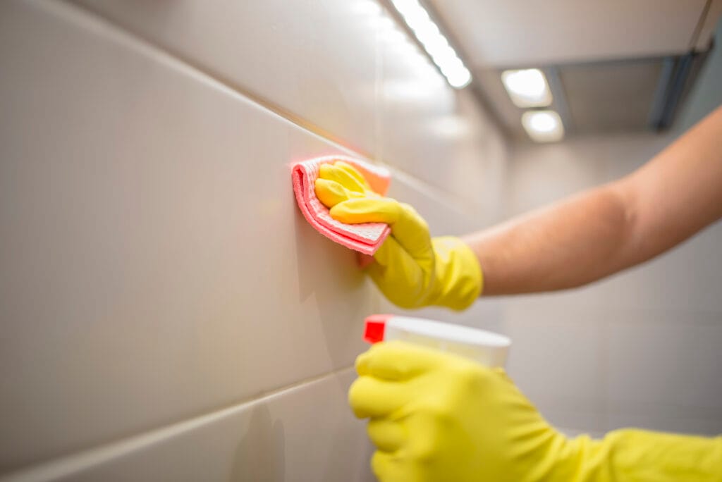 A person wearing yellow gloves cleans a white tiled wall with a pink cloth and spray bottle under bright lighting—perfect for Los Gatos move-out cleaning or property managers prepping homes.