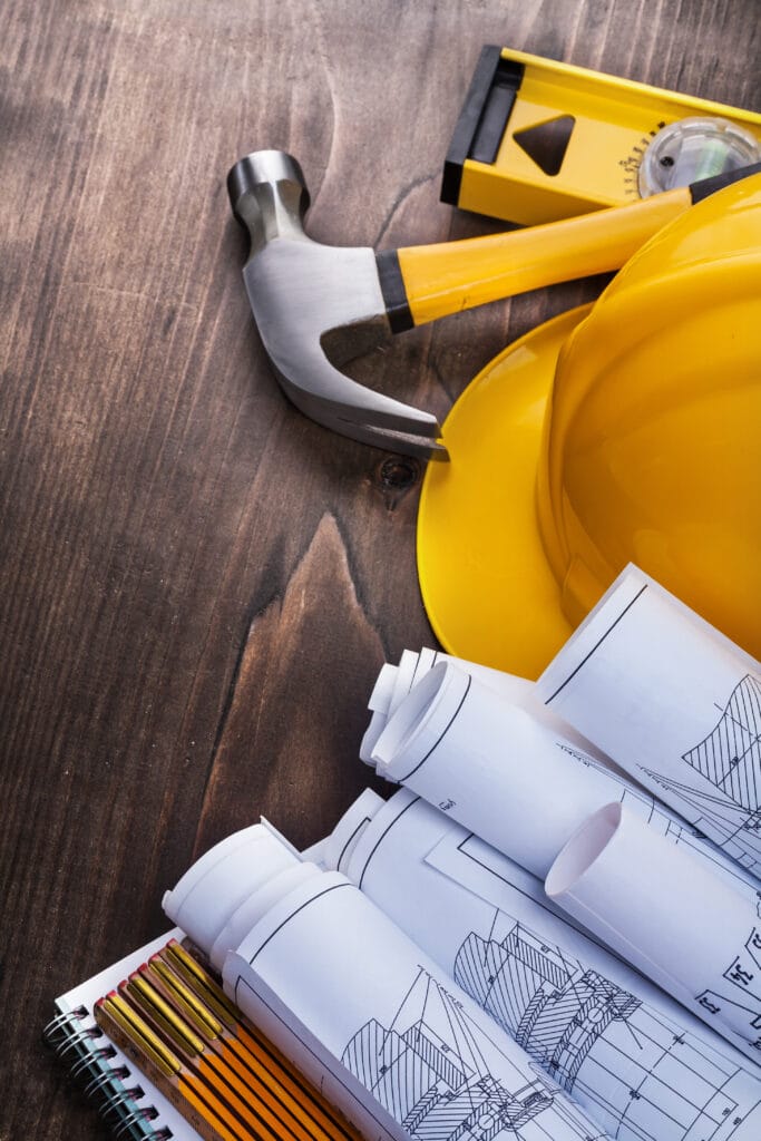 A yellow hard hat, hammer, level, rolled blueprints, pencils, and a spiral notebook are arranged on a wooden surface—ideal for Saratoga homeowners planning their next project or scheduling post-construction cleaning.