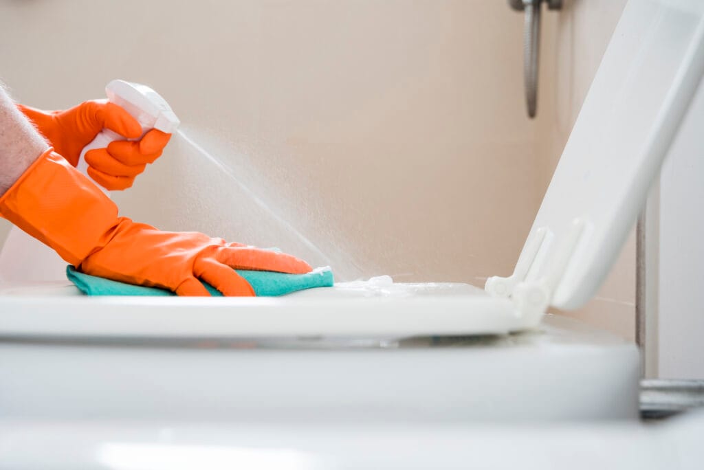 A homeowner in Saratoga wearing orange gloves sprays cleaner and wipes the lid of a white toilet with a green cloth, showcasing thorough bathroom deep cleaning.
