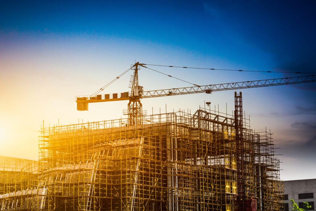 A construction site with scaffolding surrounds a building under development in Saratoga, while a tower crane stands above the structure against a blue sky at sunset, ready for post-construction cleaning.