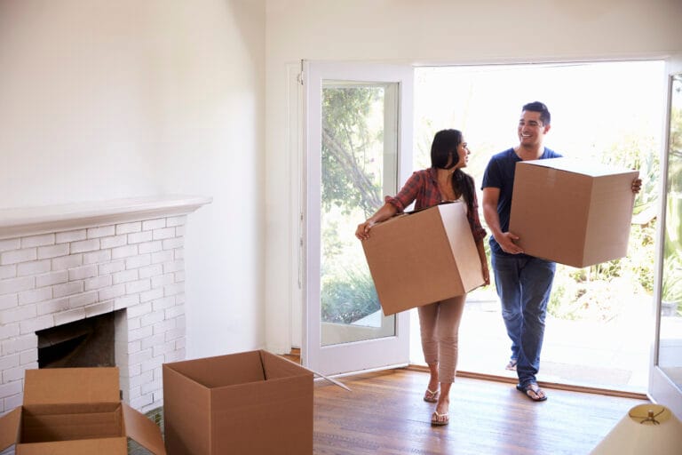 A man and woman carry cardboard boxes into an empty room with a white fireplace and open door, their spotless exit perfect for real estate agents or anyone seeking move-out cleaning in Cupertino as they settle into their new home.