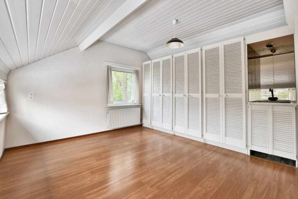 Empty attic bedroom in San Jose with wood flooring, white paneled ceiling and walls, built-in closets with louvered doors, a window, and a small sink area—ideal for Real Estate Agents or Roommate Move-Out Cleaning needs.