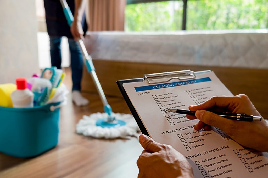 Person holding a cleaning checklist and pen in the foreground, with another mopping the floor near a bed in the background; cleaning supplies are visible in a blue bucket—perfect for Builders seeking Jobsite Ready Turnover or Final Cleaning Campbell services.