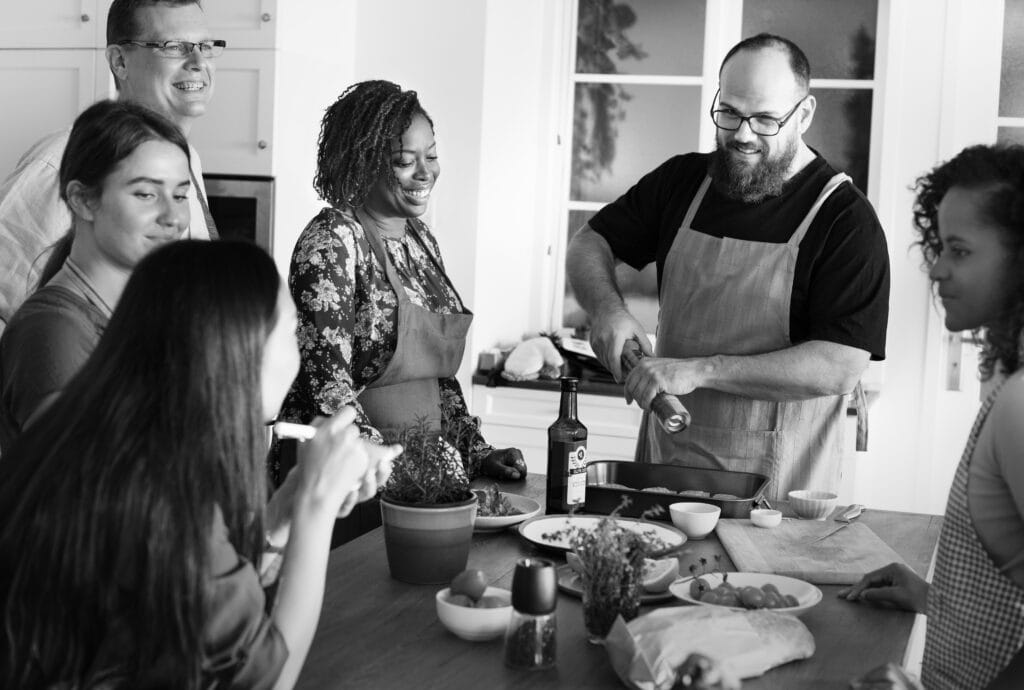 A group of six people in aprons gather around a kitchen island, preparing food and smiling together in a bright Campbell kitchen, enjoying the warmth of shared spaces and the benefits of customized kitchen cleaning.
